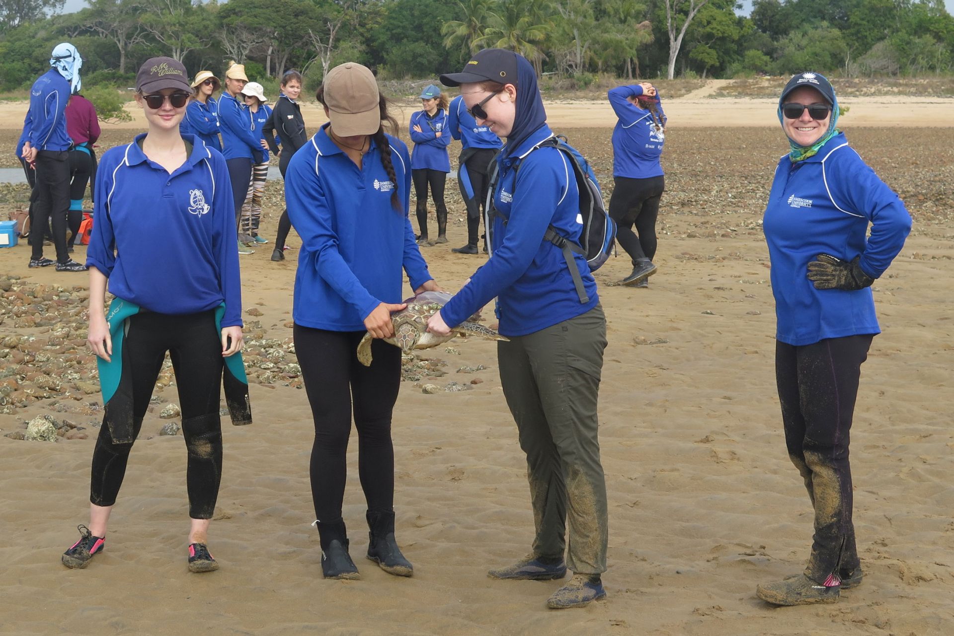 Four women wearing SunSmart clothing on a beach, two are holding a turtle between them