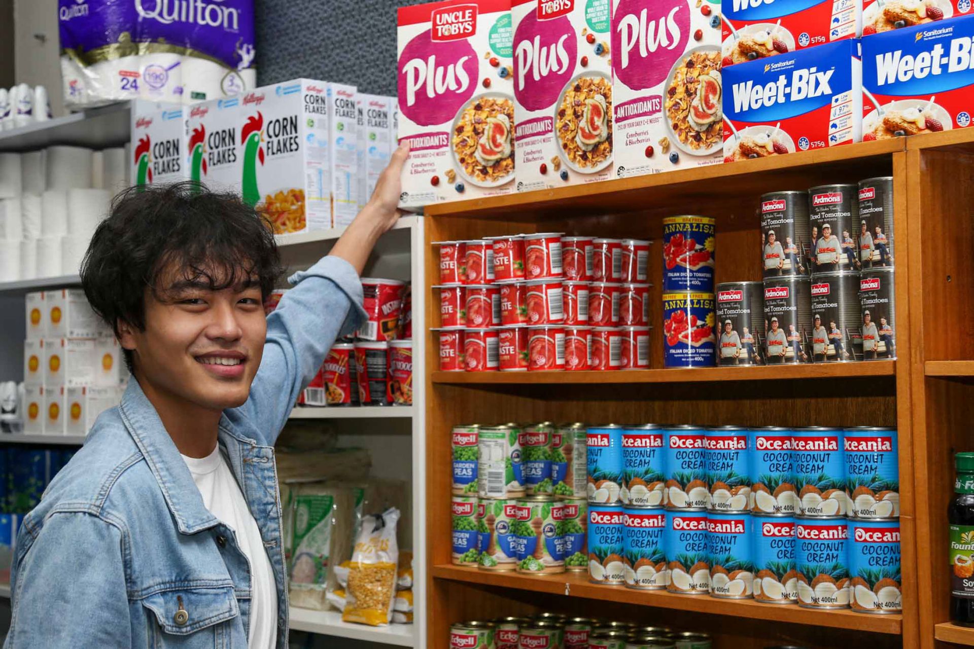 Male Nepalese student reaches up to a box of cereal on the top shelf of a fully stocked pantry