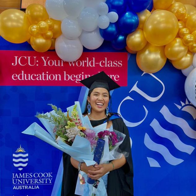 JCU student Tia at her graduation. She is posing in front of a JCU sign, holding a large bouquet of flowers. 