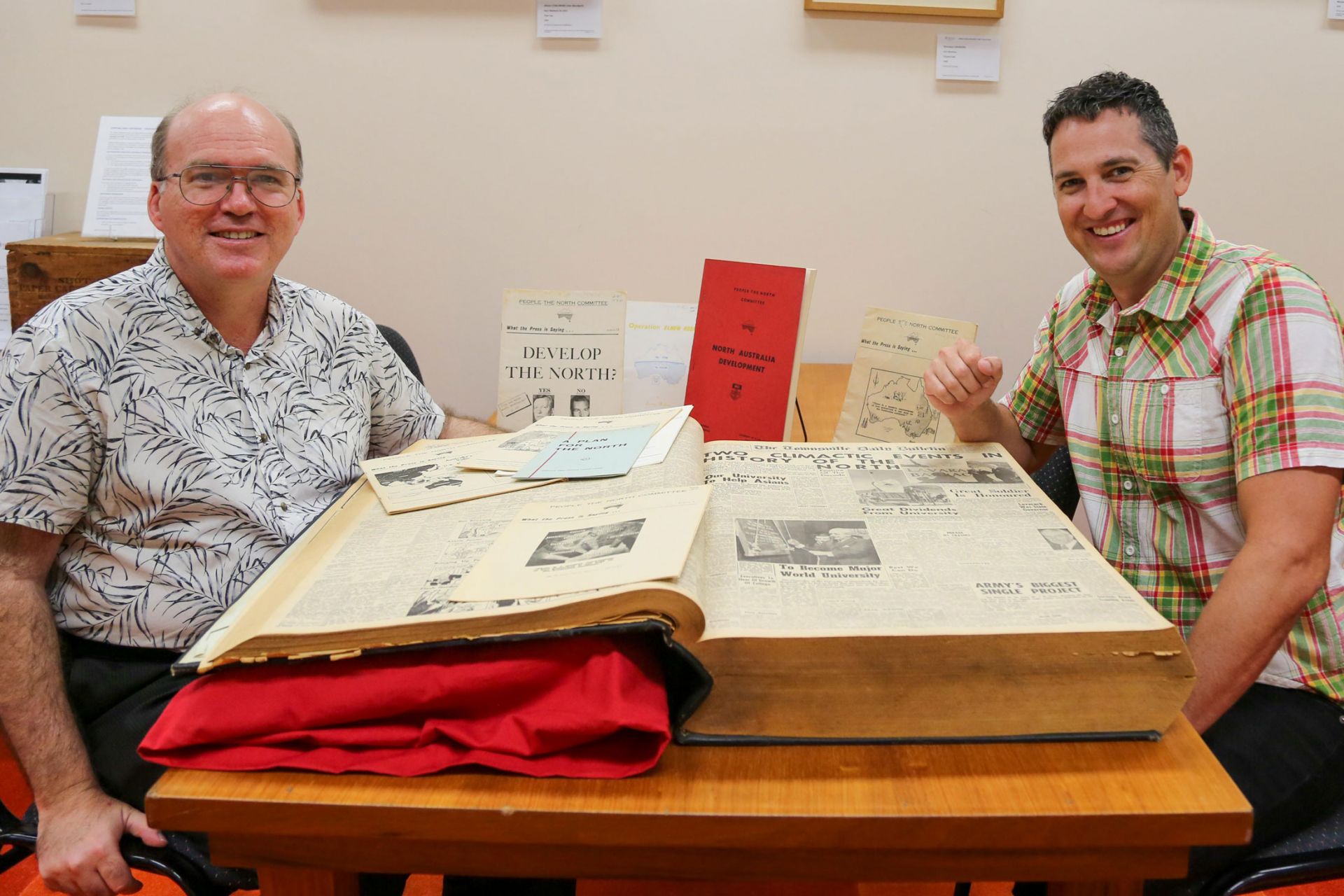 Two men, one on either side of a large bound collection of newspapers