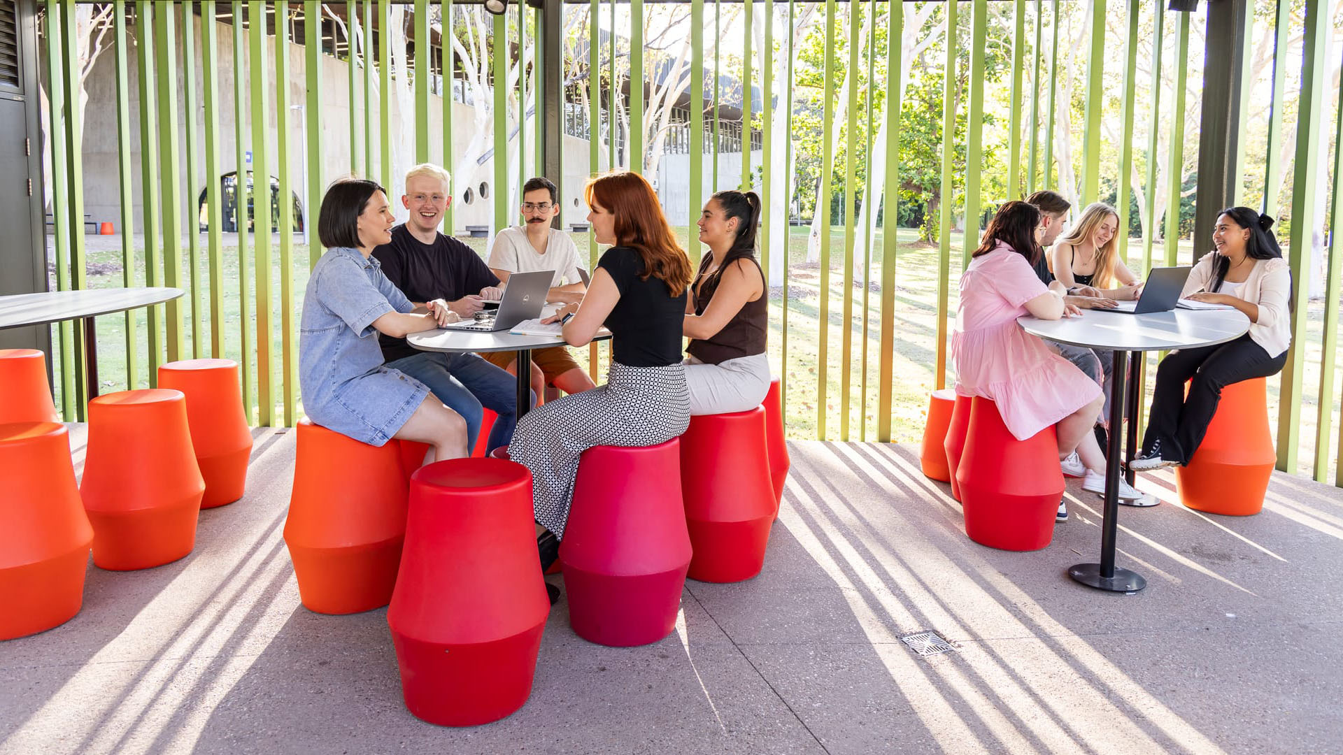 Groups of students sit at tables in front of tall green poles with sunlight casting through. 
