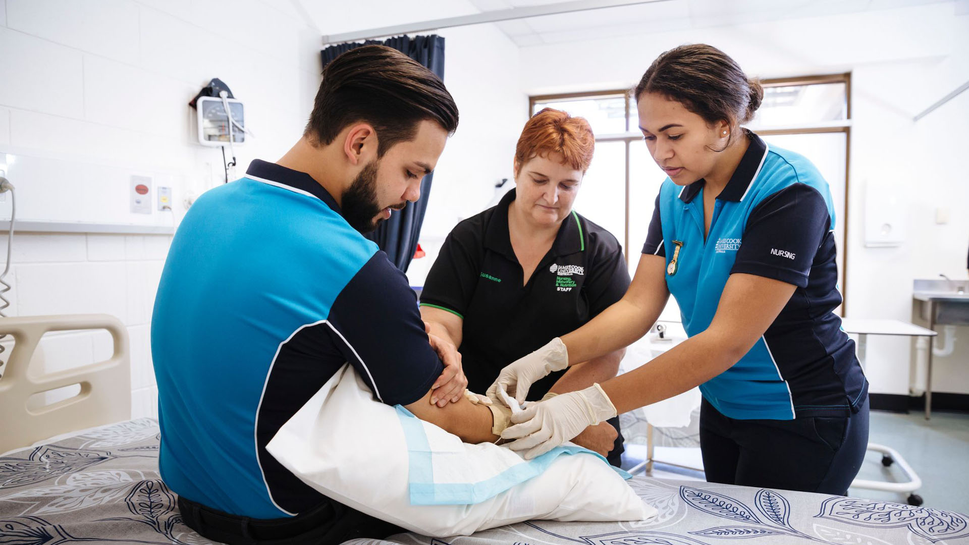 A nursing teacher helps a nursing student practice applying gauze to a fellow nursing student. 
