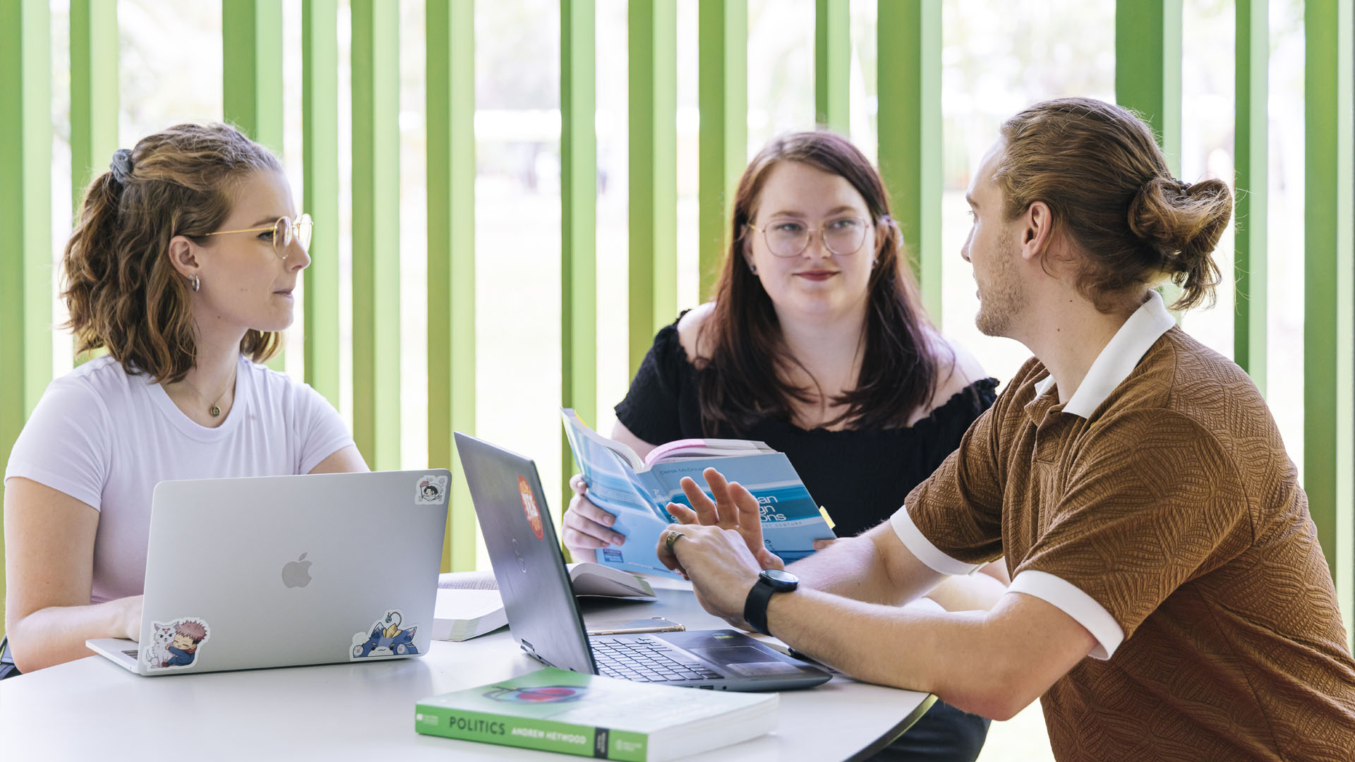 Students with laptops sit grouped around a table having a discussion. 