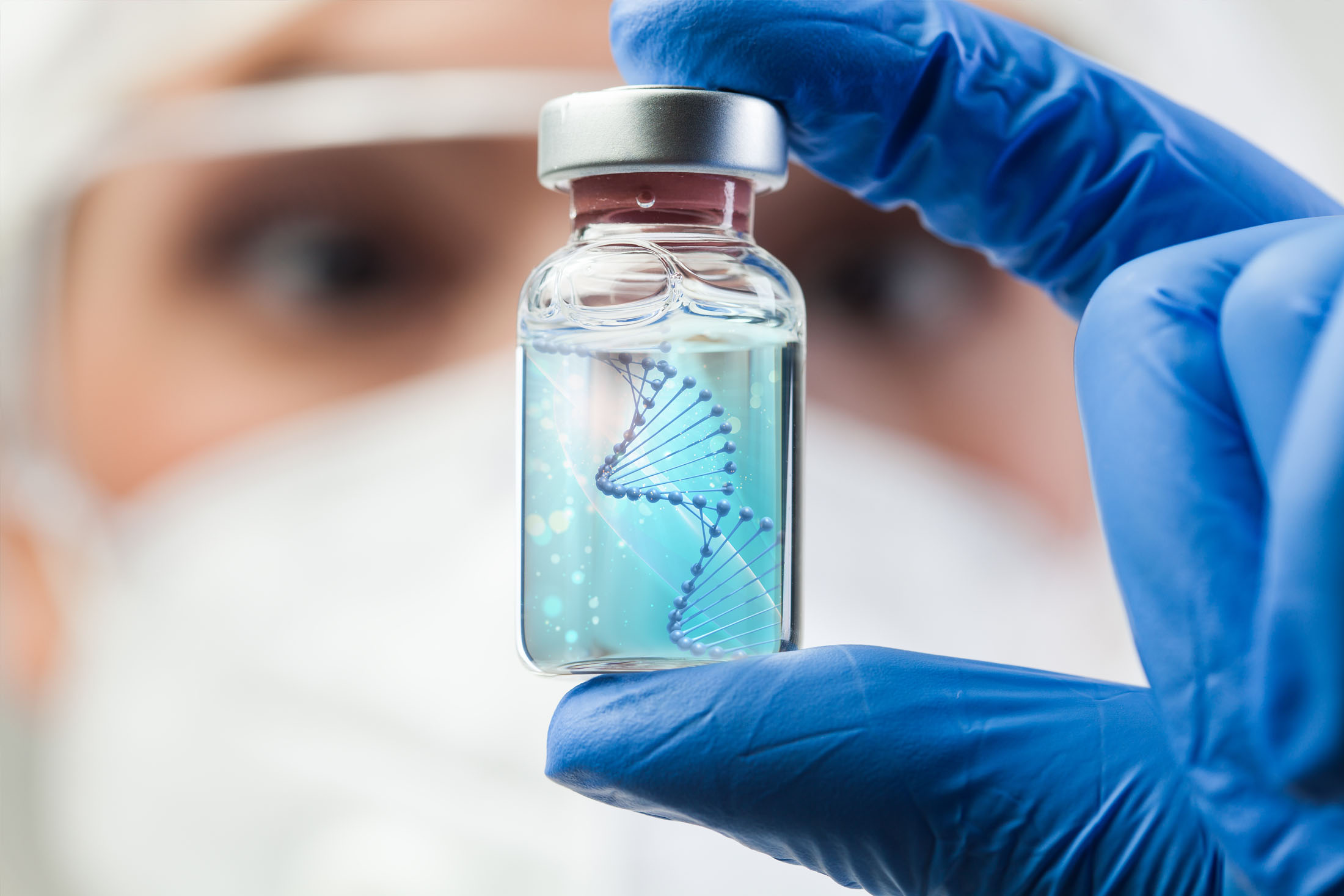 A close up of a scientist's blurred face. In front of the face is a clear bottle with a DNA helix inside, which is being held between the scientist's blue-gloved thumb and finger. 