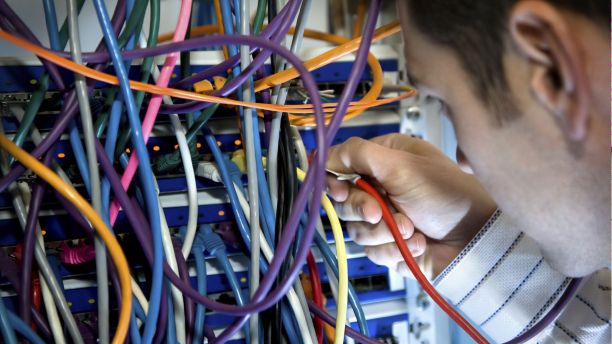 A computer server with dozens of multicoloured wires sticking out. The back of a male head is on the right hand side, positioned as if the person is looking at the computer server.