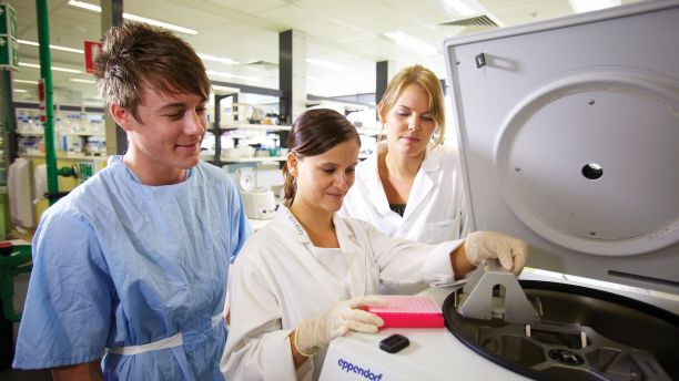 A man and two women stand next to an open centrifuge machine and are looking inside. 