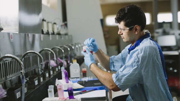 A man wearing a blue medical lab coat and safety goggles sits at a stainless steel bench holding a sample container. 