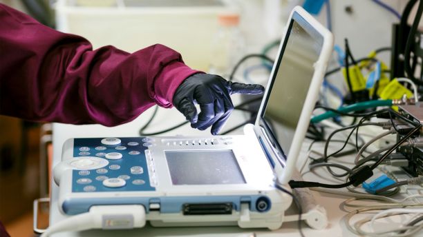 A gloved hand points to a laptop with many cords coming out of it. 