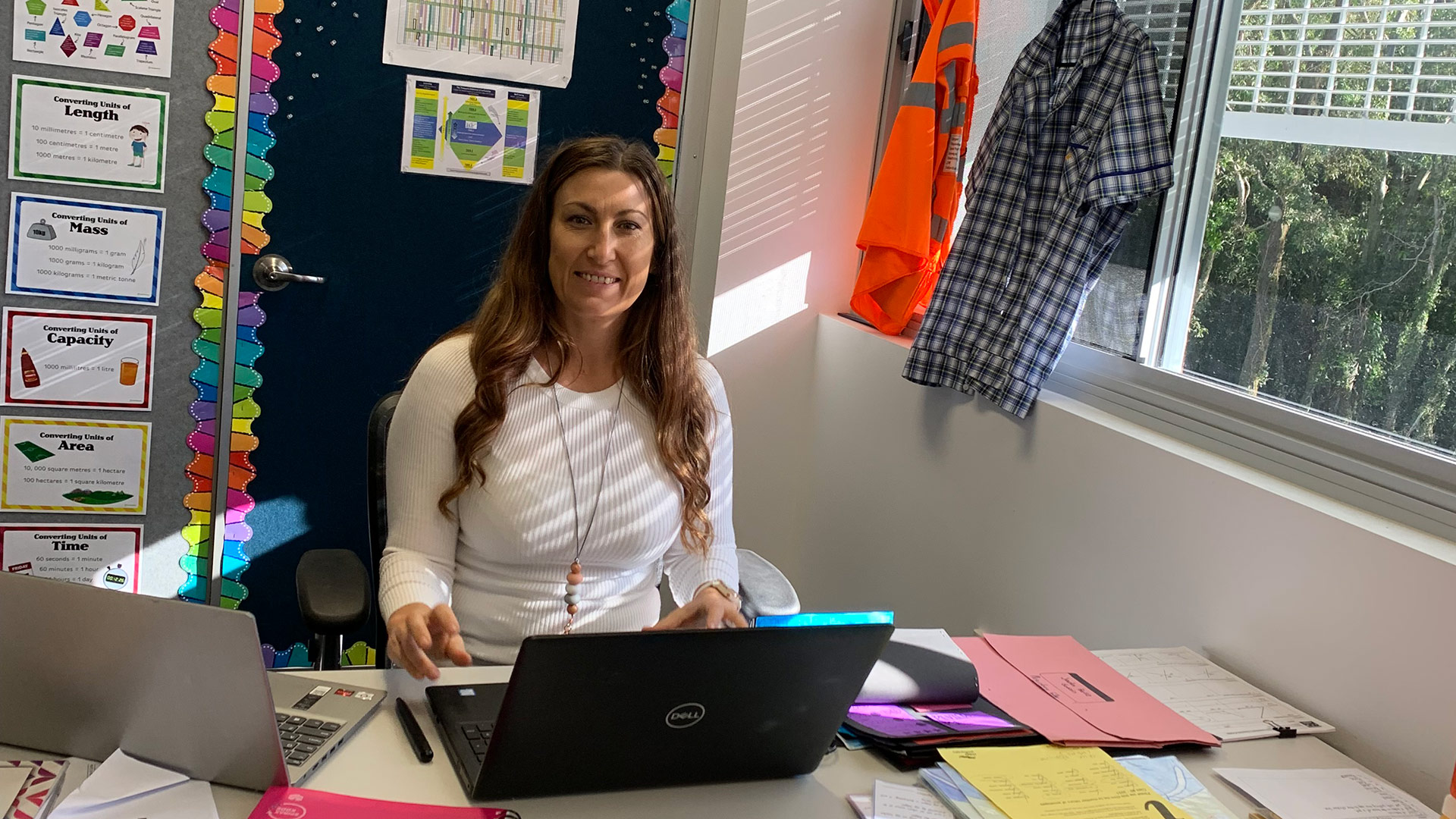 Year Six teaching Courtney Hawkins sits at her desk smiling in front of a laptop with classroom posters on the wall behind her. 