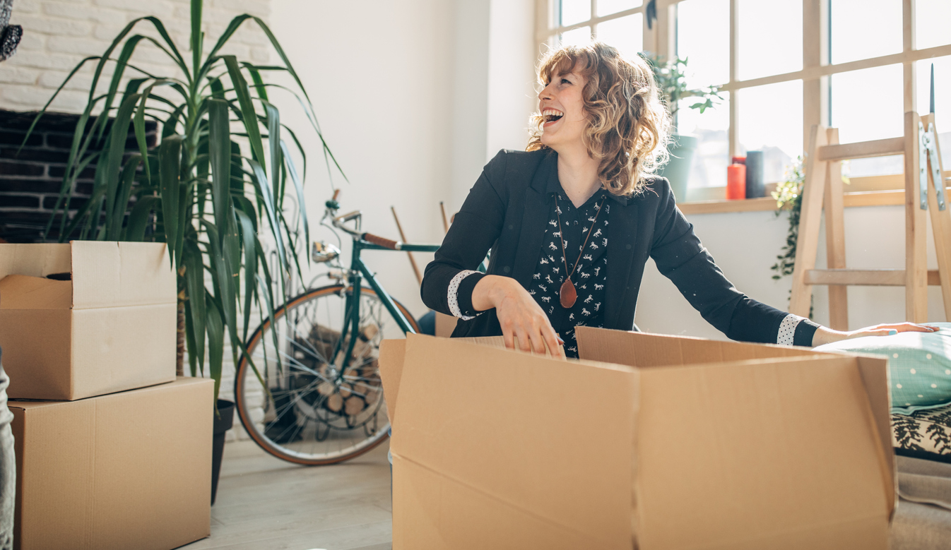 A smiling woman is laughing while reaching into a packing box and is surrounded by other packing boxes. 