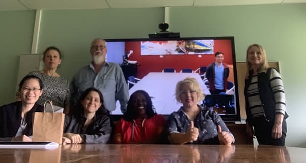 An Assortment of 7 people standing infront of a Screen showing a video call with an additional person, all posing to the camera.