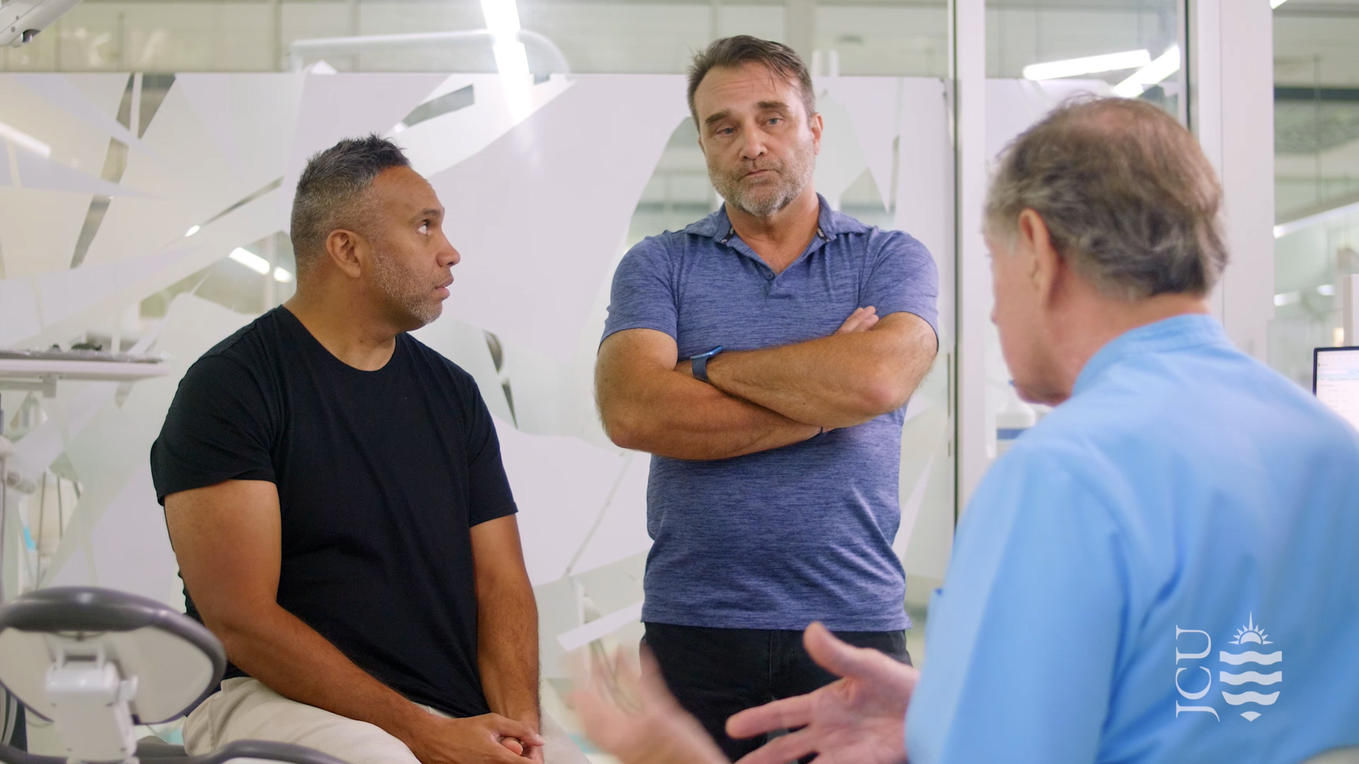 man standing with arms crossed while dentist talks to patient. 