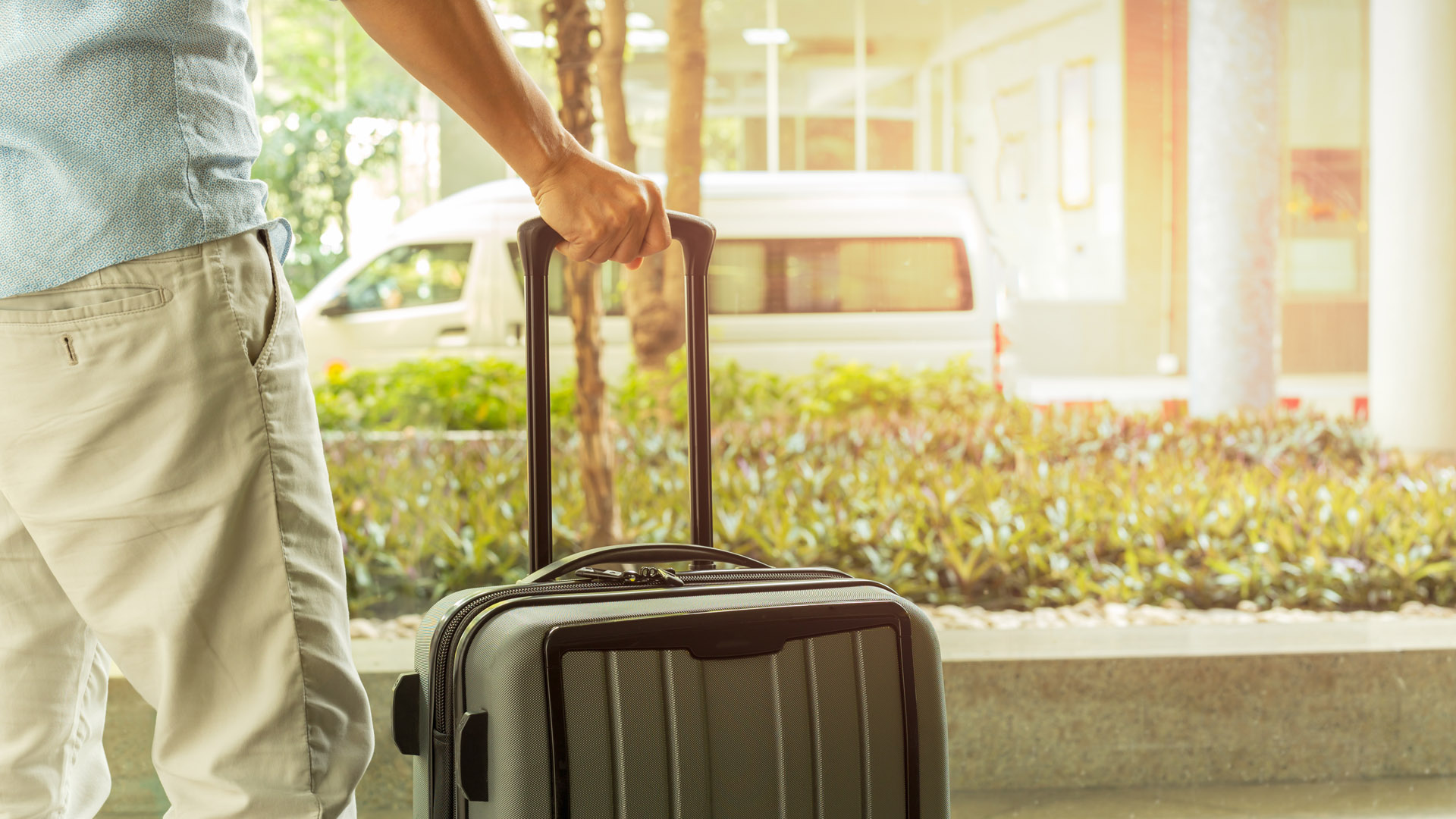 A man stands at an airport, faceing away from the camera and holding a suitcase. 