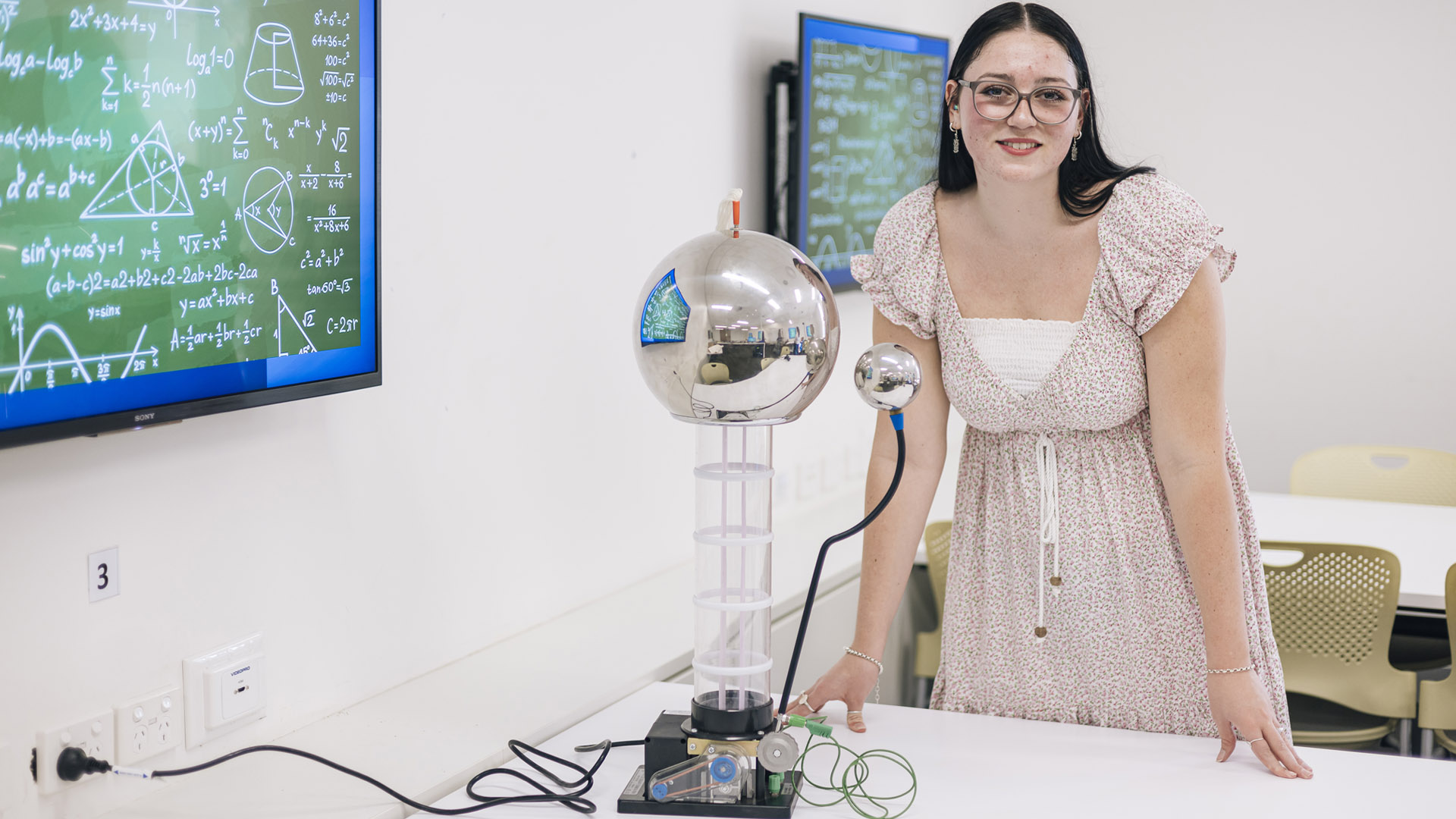 A JCU Education student stands next to a scientific instrument with math equations on the board behind her. 