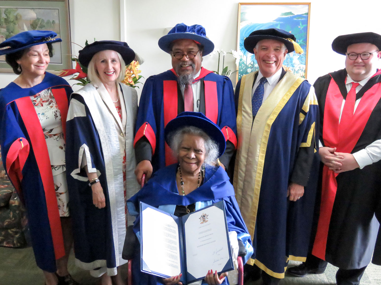 JCU academics in robes stand Behind Bonita Mabo, who is holding her honorary doctorate and wearing academic dress