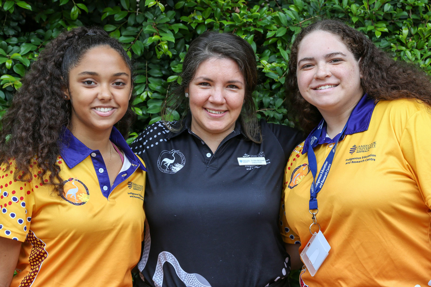 Two female students wearing yellow/orange Winter School shirts stand on either side of a female teacher wearing a black IERC shirt. 