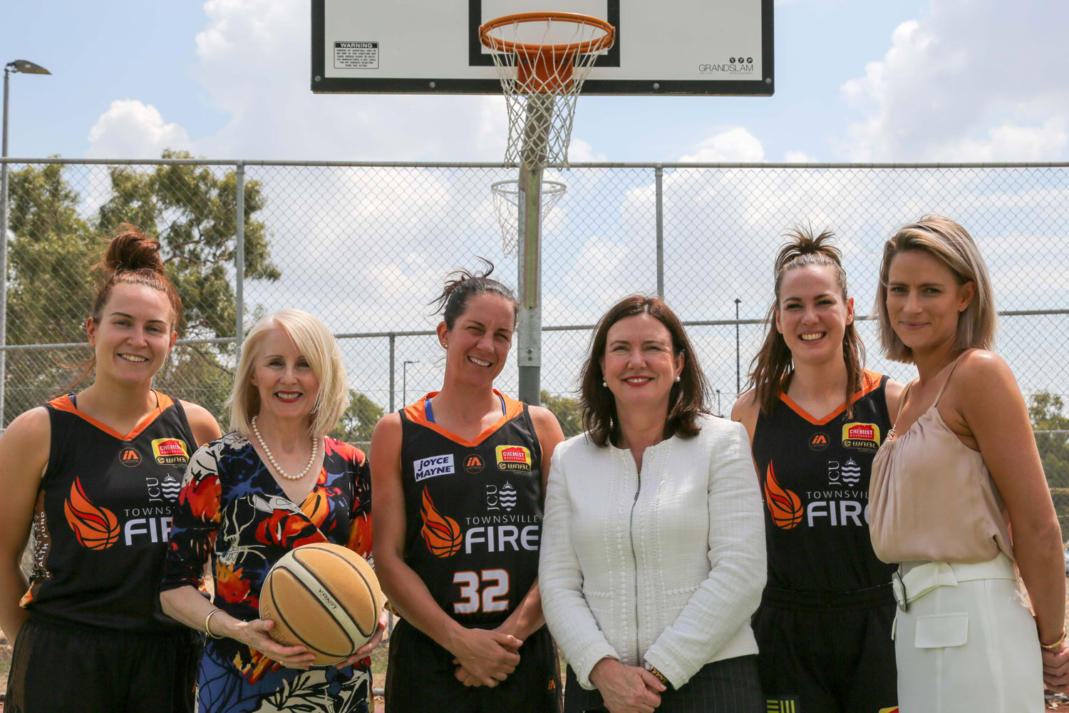 Six women, three wearing basketball uniforms and three wearing office attire stand in a line in front of a basketball hoop