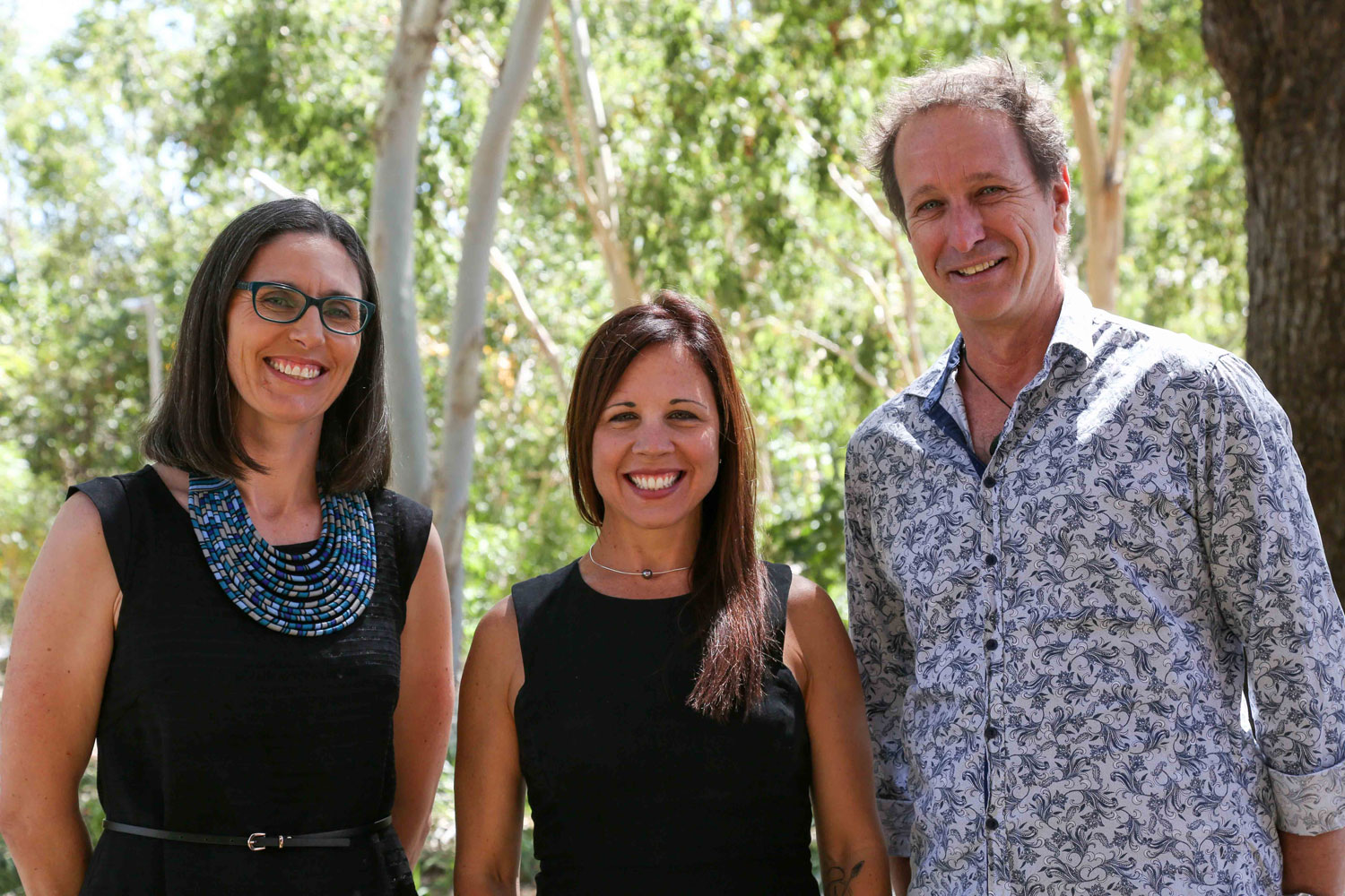 Two women and a man wearing casual clothes and standing outside underneath trees stand together and smile at the camera 