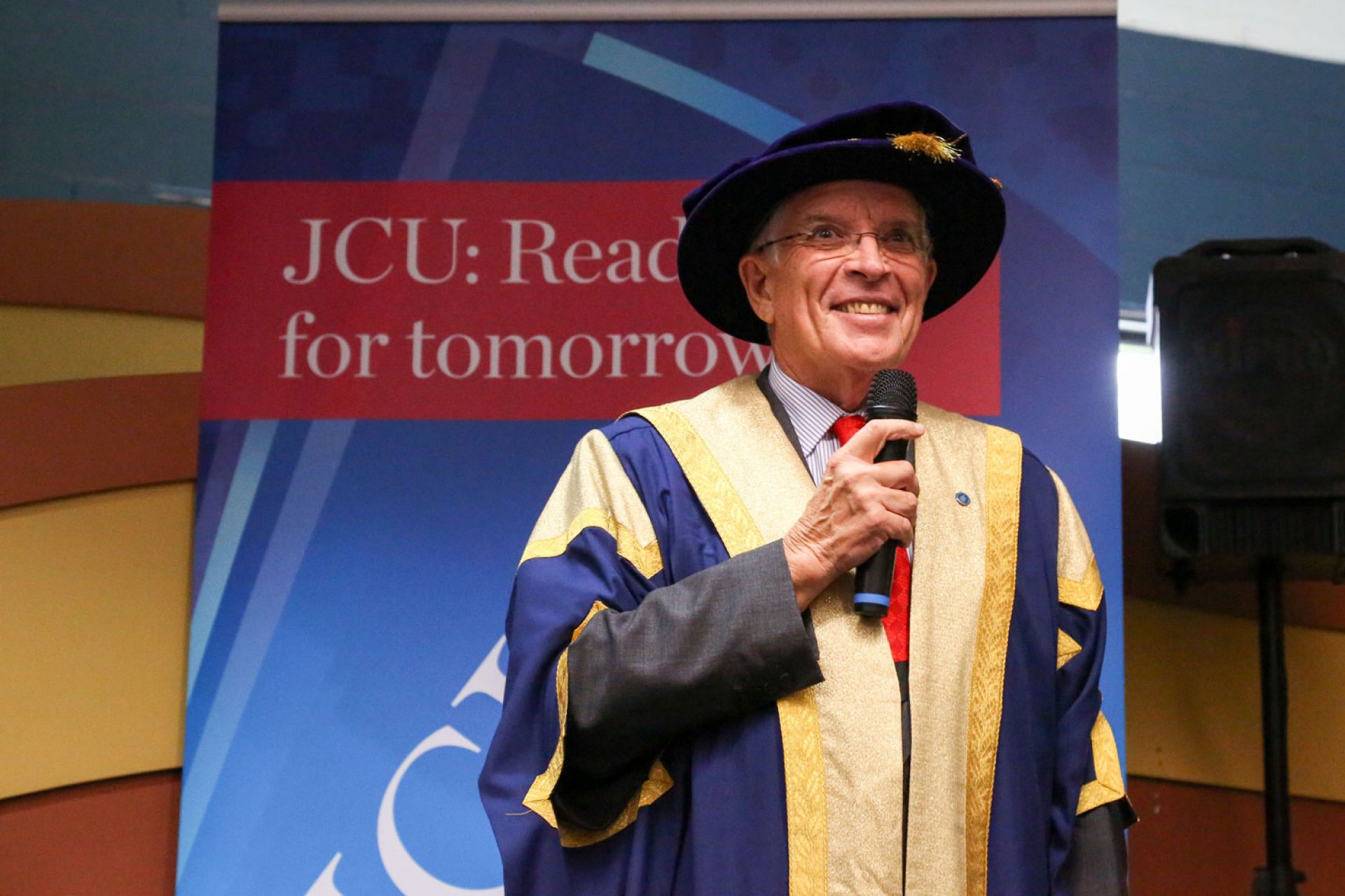 Bill Tweddell smiles while speaking into a microphone, wearing his chancellor regalia, in front of a JCU banner