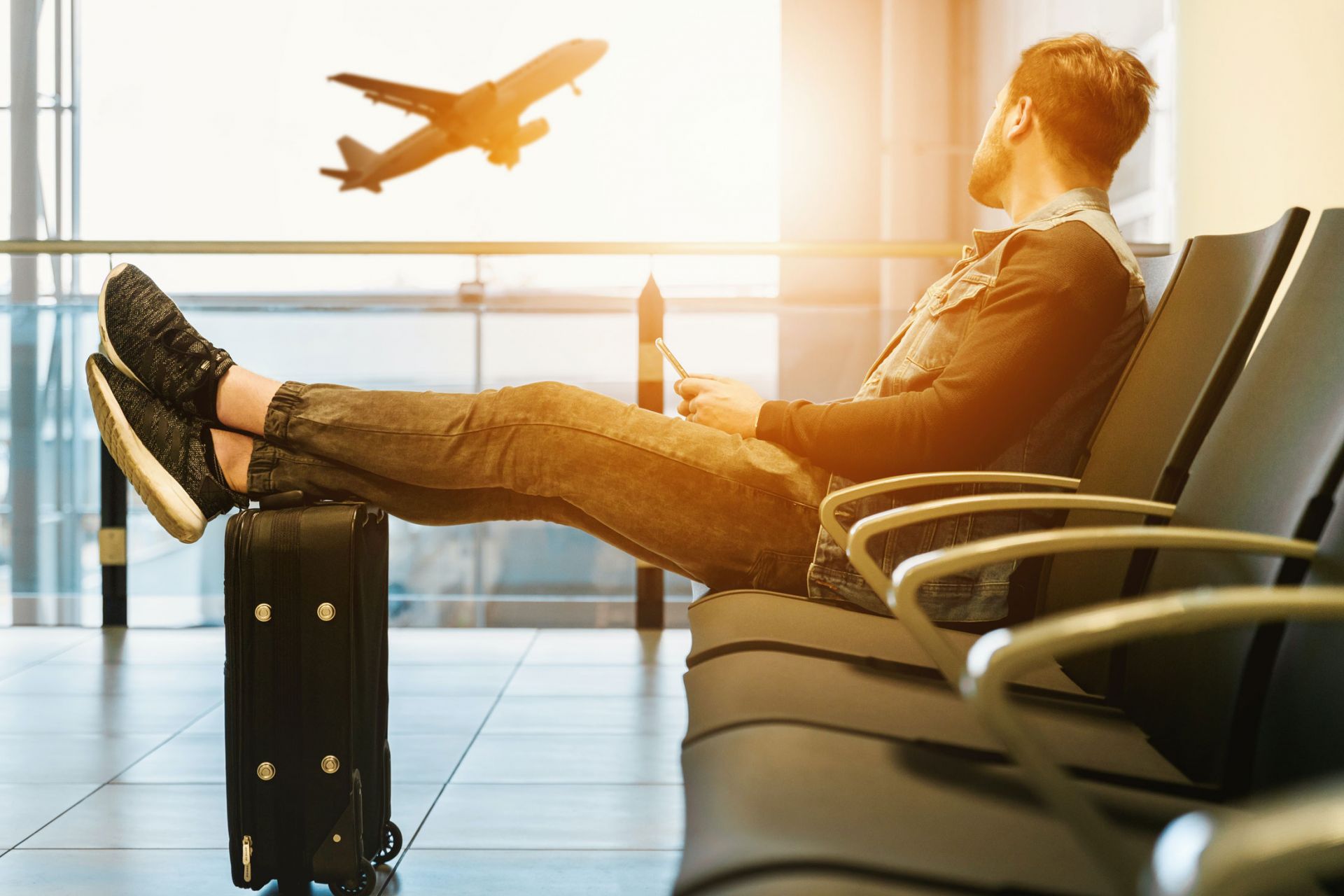 man sitting in an airport departure lounge, looking out the window to a plane taking off