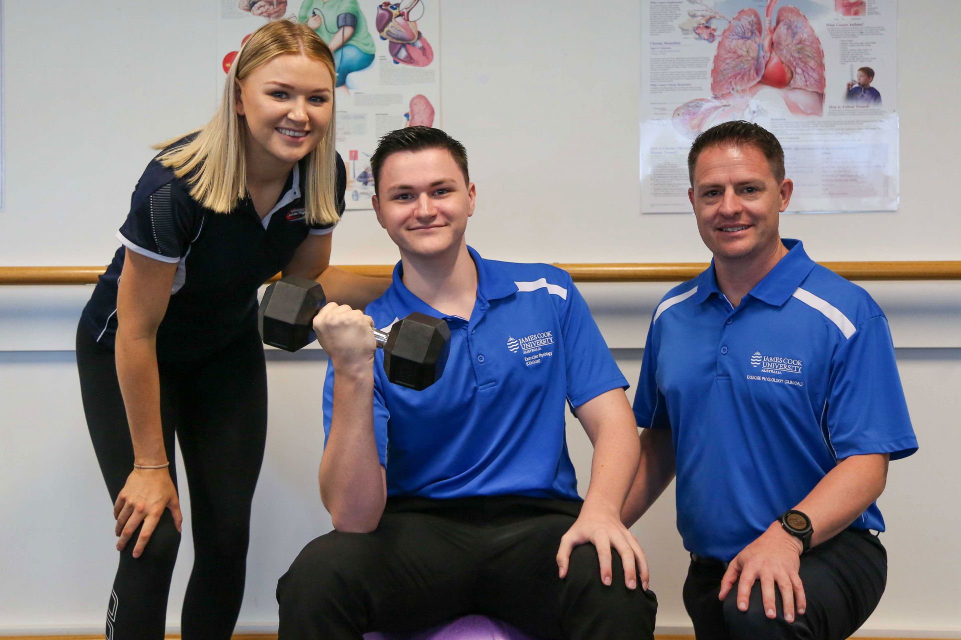 Young man sitting on a purple fit ball doing a bicep curl, with a young woman to one side, and a man to the other 