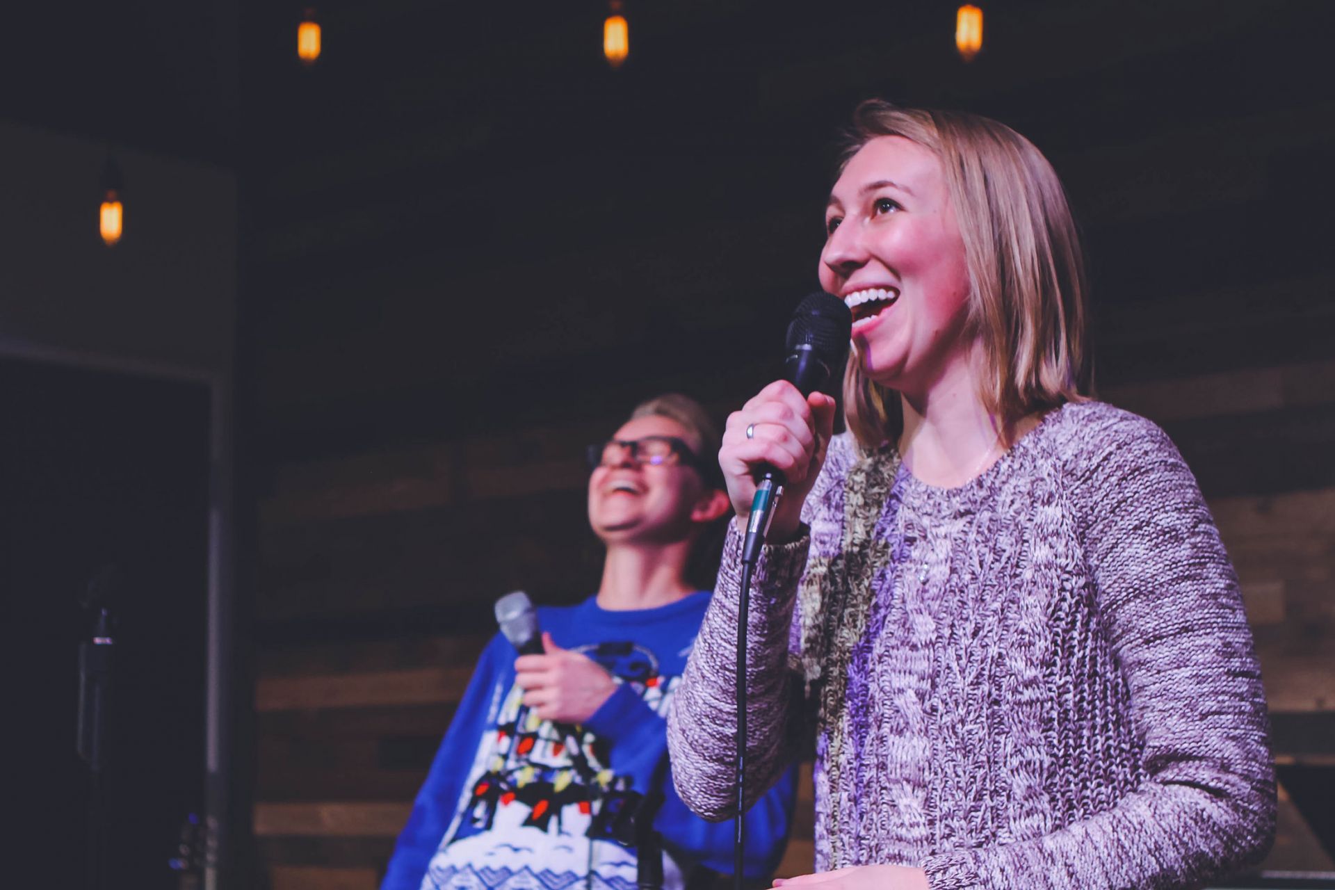 woman in focus holding a microphone singing, behind and out of focus is a man holding a microphone