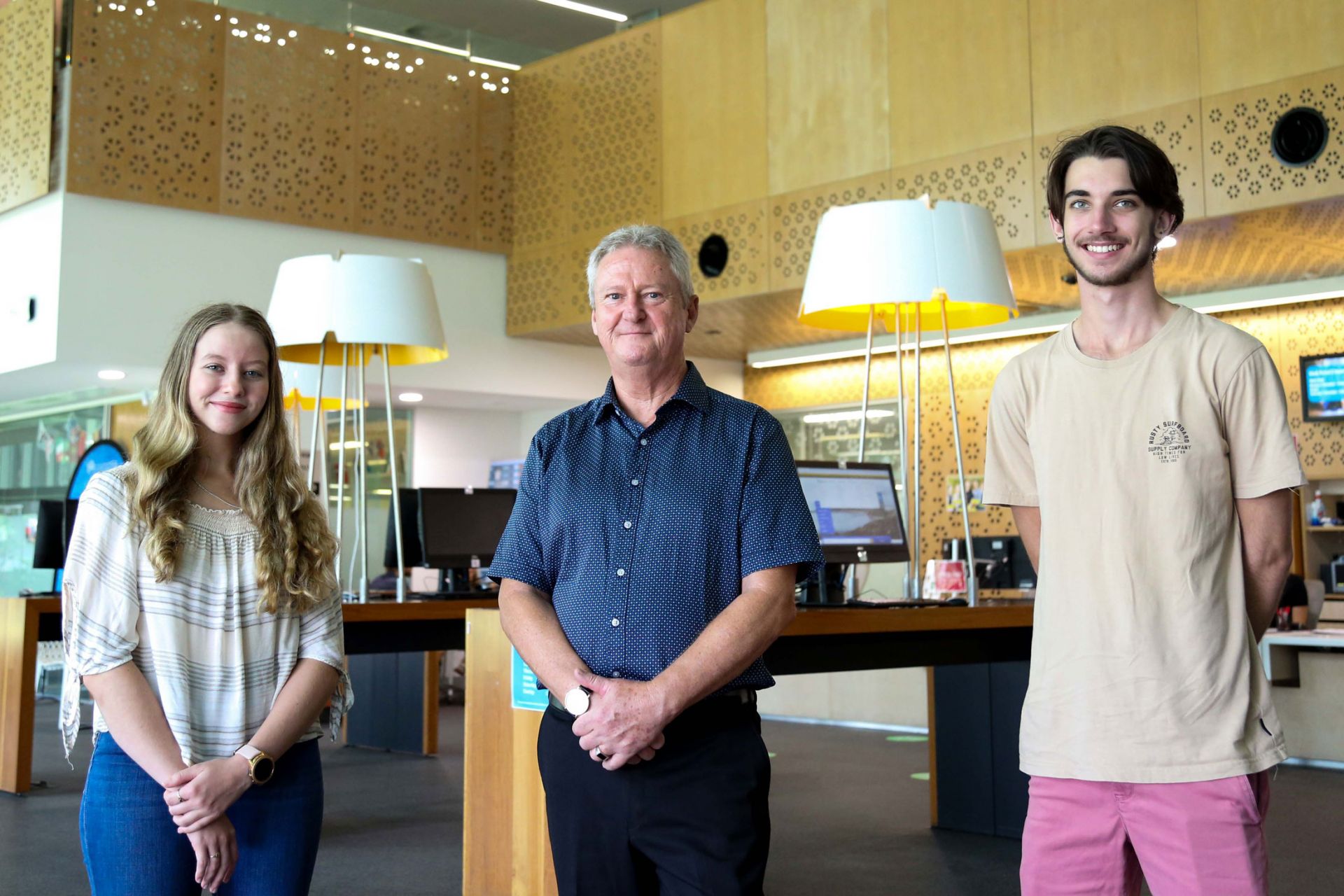 A young woman and a young man stand on either side of an older man inside the foyer of a university building