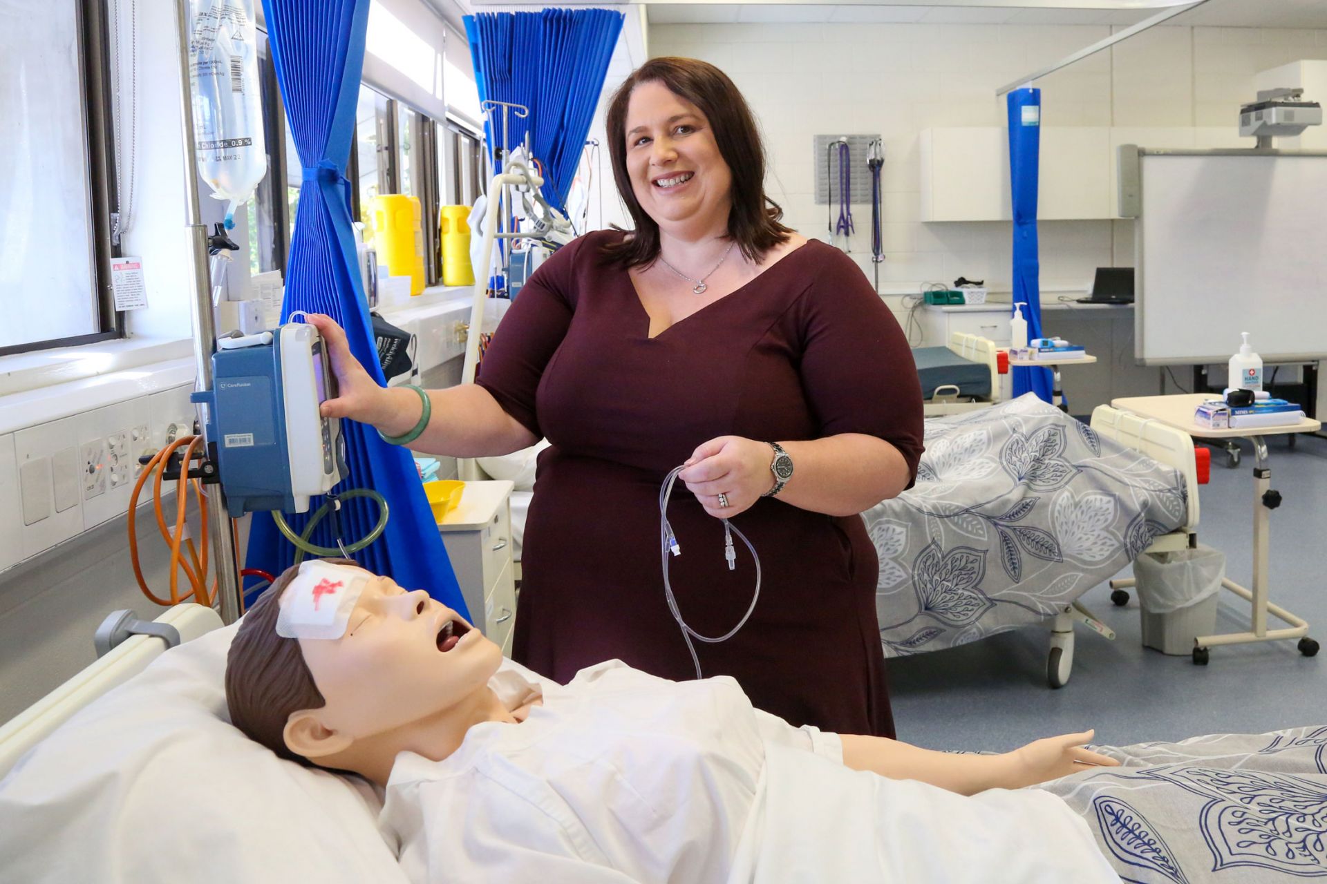 Tanya stands next to a university lab hospital bed with a mannequin patient in it.