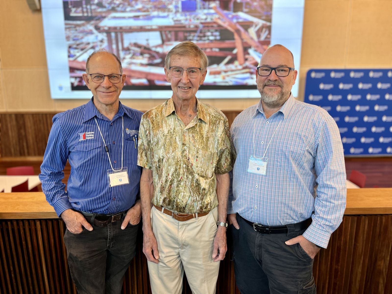Three team members of the Cyclone Testing Station stand in front of the photo of destruction following Cyclone Tracy