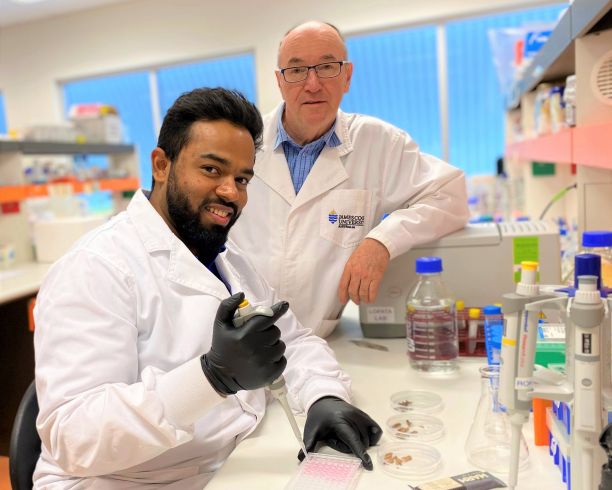 JCU PhD student Shay Karnaneedi smiling and sitting at a lab bench with Professor Andreas Lopata standing behind him. 