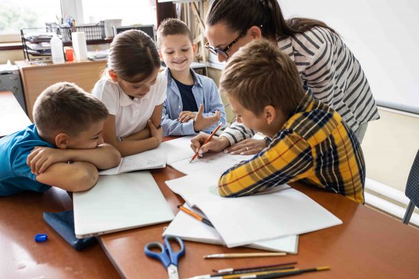 Teacher leaning over at a desk helping four young students surrounded by notebooks and pencils