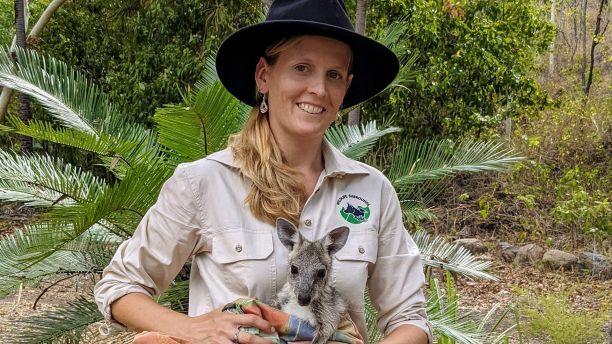 JCU Alumni Shakira Todd smiling while holding a young wallaby in a fabric pouch in her arms. 