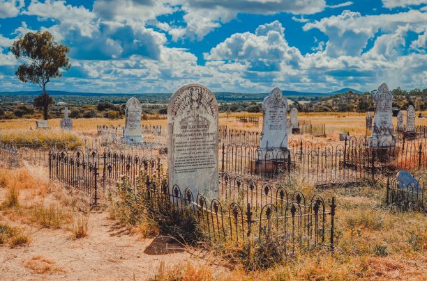Large historic headstones in a cemetery with sand and dried grass around and bright blue skies and clouds in the background. 