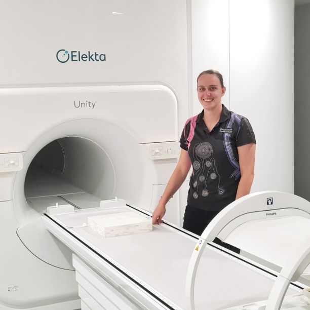 JCU Alumni Ari Brodmann smiling and standing next to the MR-Linac machine that provides targeted radiation treatment.