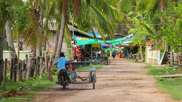 Two children driving down a street in Laos surrounded by palm trees with buildings in the background. 
