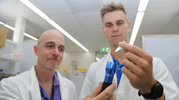 Associate Professor Patrick Schaeffer on left and PhD Candidate Casey Toft on right, each wearing lab coats and holding up small vials. 