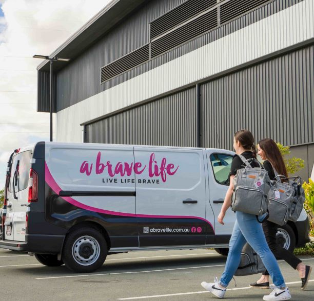 Two women smiling and carrying grey backpacks walk towards a black pink and white van with a brave life written across the side. 