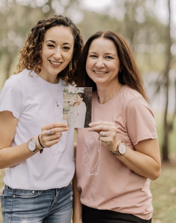 JCU Alumni Melissa Redsell and her daughter standing next to each other smiling and holding up a small photo of when they were younger. 