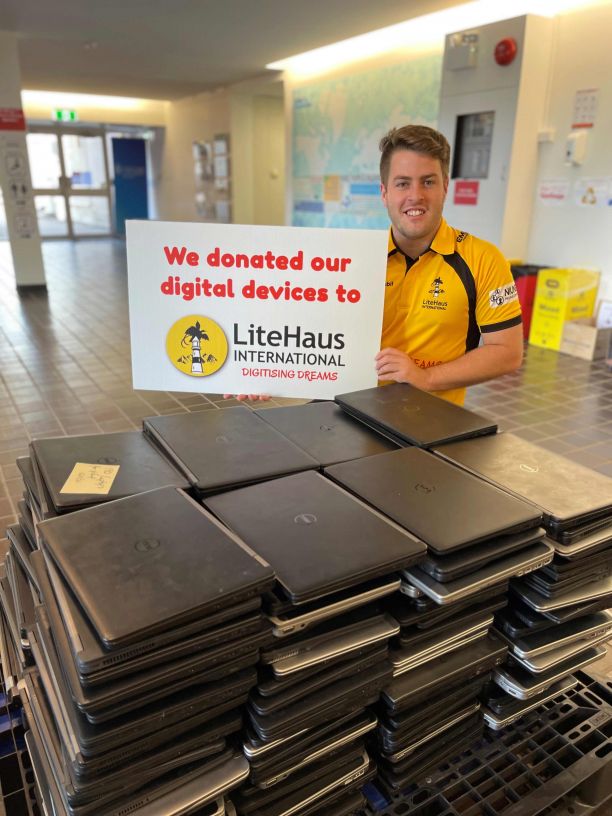 JCU Alumni Jack Growden holds up a sign saying 'we donated our digital devices to LiteHaus International' while he smiles standing behind a large pile of laptops on a desk.