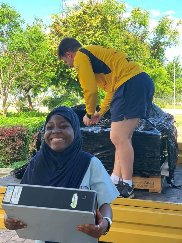 JCU Alumni Jack Growden stands on the tray of a ute unpacking computers from a pallet while a school student smiles holding one of the computers.