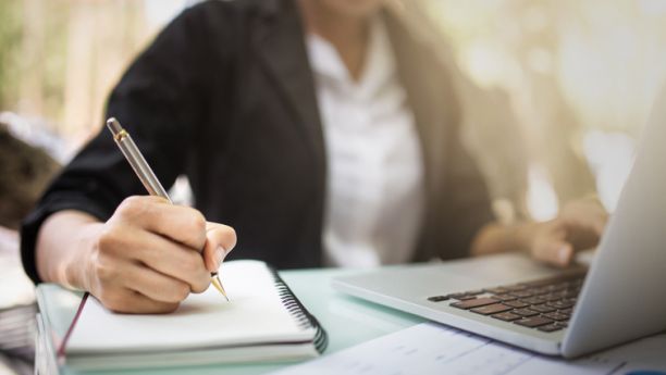 A person wearing a white shirt and black jacket sits at a desk writing with a pen in one hand while looking at a laptop. 