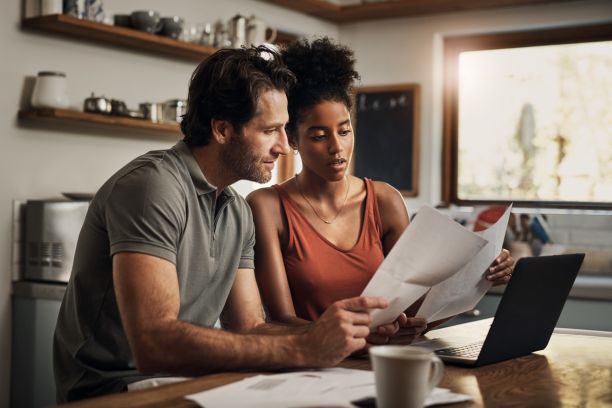 A man and a woman sit at a kitchen table with a laptop while each holding up pieces of paper and looking at them together. 